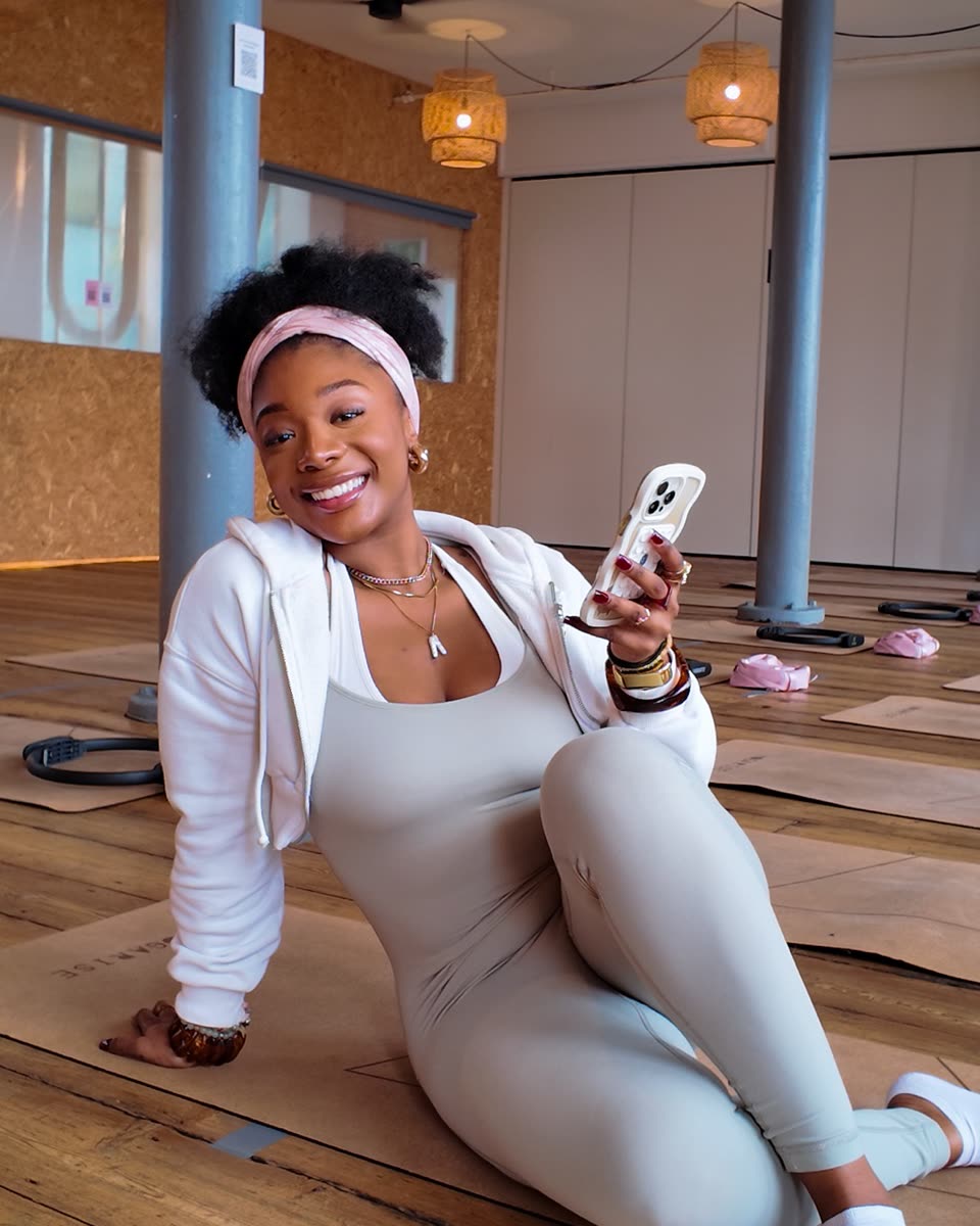Steph laughing on the floor of a sunny studio, in a pink headband and white jacket, holding her phone