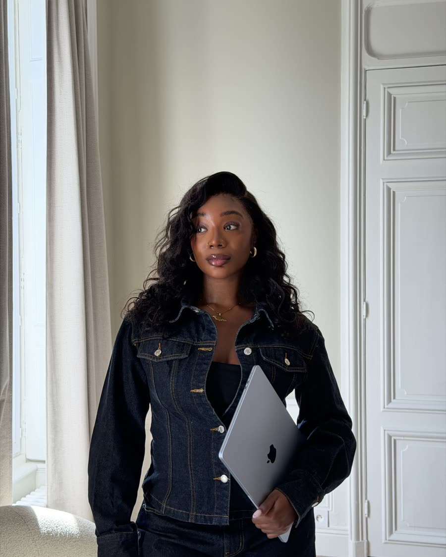 Steph in a dark denim jacket and jeans, holding a laptop in a bright room with tall windows