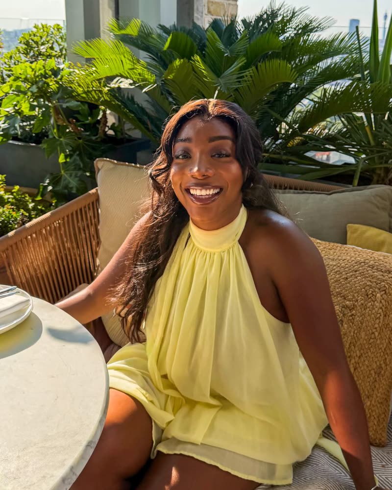 Steph smiling on a sunny rooftop terrace in a yellow dress, surrounded by plants